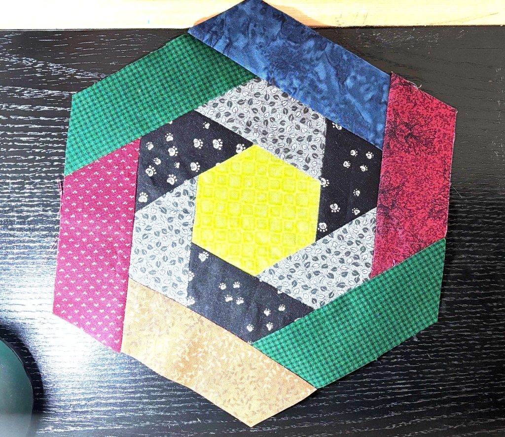 still image of the quilt block from the animation. the center is a green hexagon. alternating grey and black trapezoids wrap around that green hexagon. the outer ring alternates between red, green, and blue trapezoids