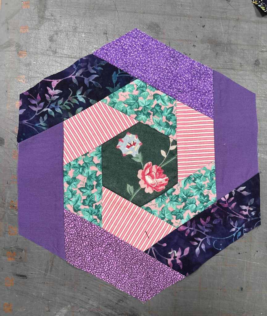 hexagon log cabin quilt block with a purple ring on the outside, pinks on the middle ring, and a rose and morning glory in the center
